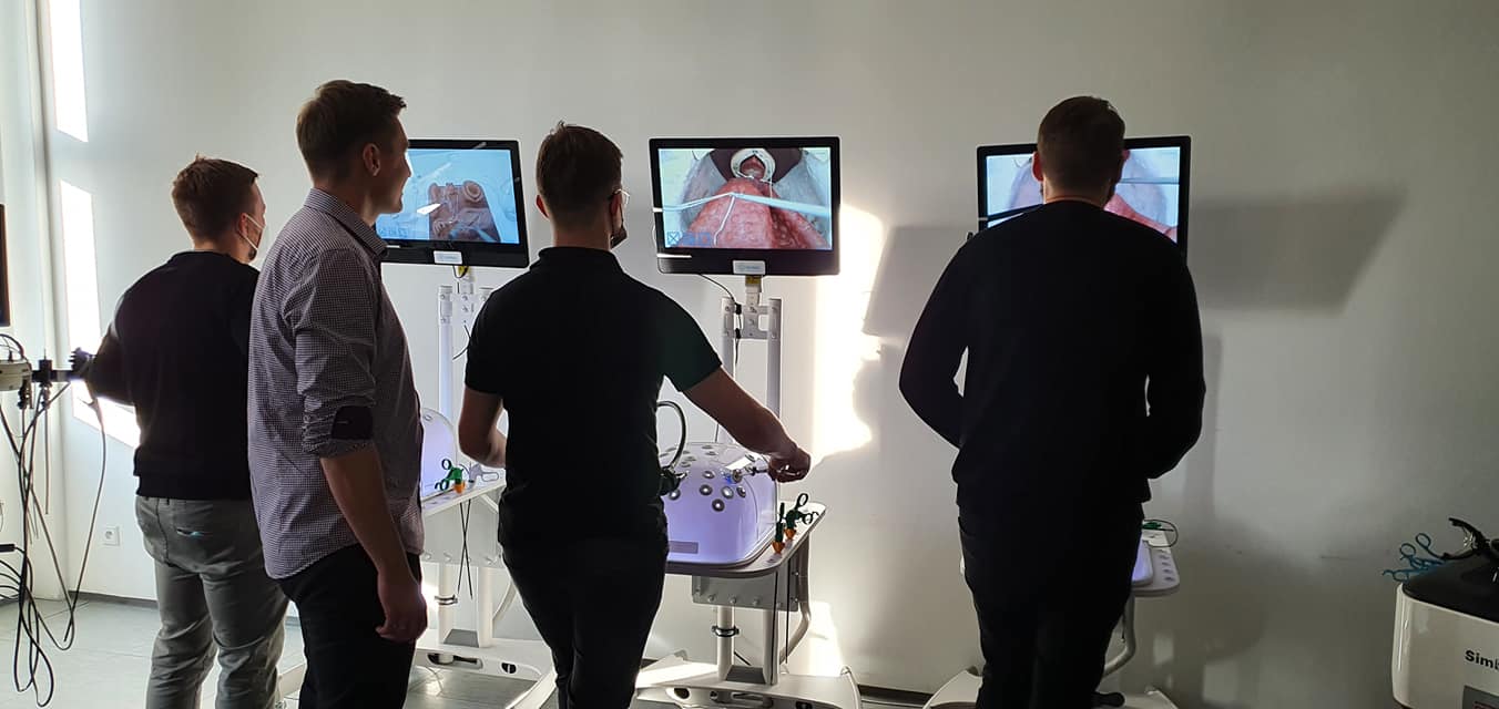  na sali szkoleniowej stoją 4 osoby odwrócone tyłem przed symulatorami medycznymi wykonując procedurę chirurgiczną uwidocznioną na ekranach monitorów